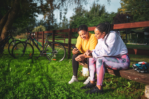 African American young woman showing smart phone to female friend while relaxing on bench in park with bicycles parked on grassy field