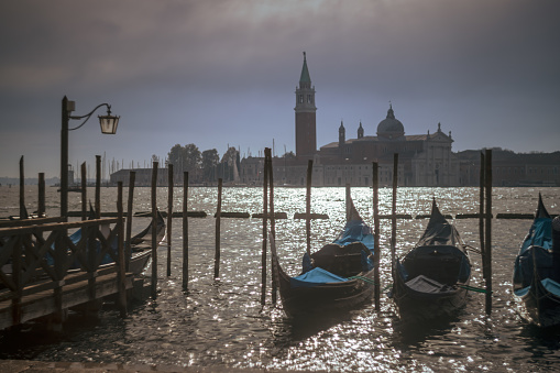 Gondolas and wooden posts in Grand Canal by pier with Church of San Giorgio Maggiore in the background against cloudy sky at Venice,Italy