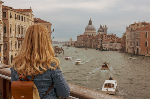 Rear view of female tourist with blond hair looking at Santa Maria della Salute and residential buildings while standing on footbridge over Grand Canal in Venice,Italy