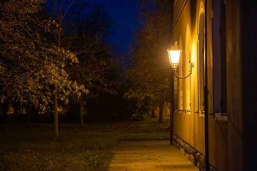 Illuminated retro electric lamp on wall of old residential building and trees growing in yard at night