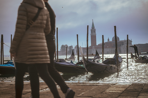 People walking on pier and gondolas with wooden posts in Grand Canal against Church of San Giorgio Maggiore under cloudy sky at Venice,Italy