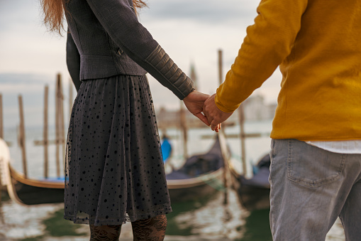 Midsection of romantic couple holding hands and standing in front of gondolas moored on Grand Canal while enjoying vacation at Venice,Italy