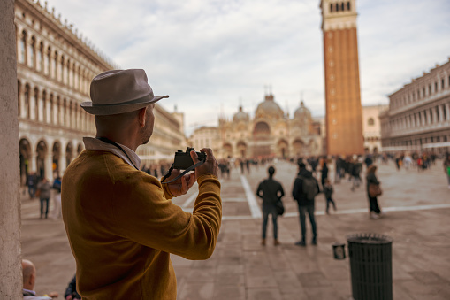 Side view of male tourist wearing hat photographing Campanile tower and Saint Mark's Basilica with camera while standing at Piazza San Marco square in Venice,Italy