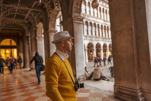 Side view of male tourist wearing hat with camera looking at Piazza San Marco square while standing in Procuratie Vecchie at Venice,Italy