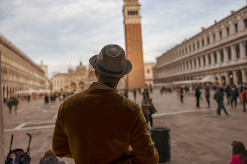 Rear view of man wearing hat looking at Campanile tower and Saint Mark's Basilica while exploring Piazza San Marco square in Venice,Italy