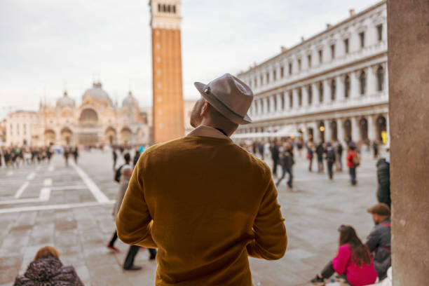 Rear view of male tourist wearing hat looking at Campanile tower and Saint Mark's Basilica while standing at Piazza San Marco square in Venice,Italy