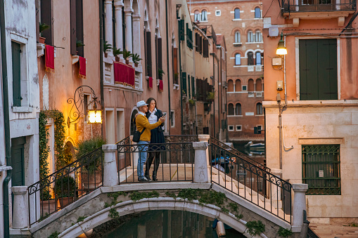 Couple using mobile phone while standing on arch footbridge over Grand Canal against historic buildings in Venice,Italy during vacation at sunset