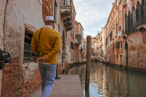 Rear view of male tourist wearing hat hiding rose behind back while walking on pier by Grand Canal between historic residential buildings in Venice,Italy