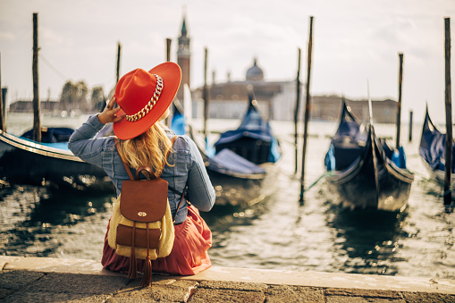 Rear view of female tourist with backpack wearing hat and looking at gondolas moored on Grand Canal and Church of San Giorgio Maggiore against sky at Venice