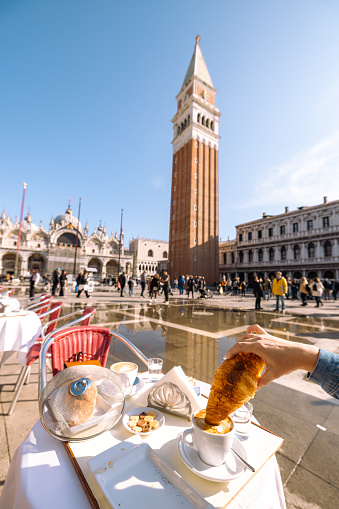 Cropped hand of man having croissant with coffee at sidewalk cafe in front of Saint Mark's Basilica and Campanile against clear sky in Venice,Italy