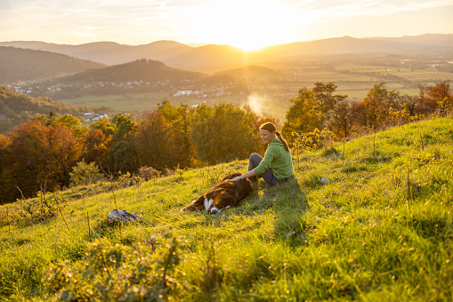 Female tourist stroking Bernese Mountain Dog lying on grassy mountain against scenic view of landscape and sky during vacation