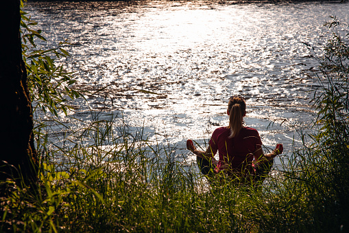 Woman sitting and practicing yoga at river bank in forest during summer