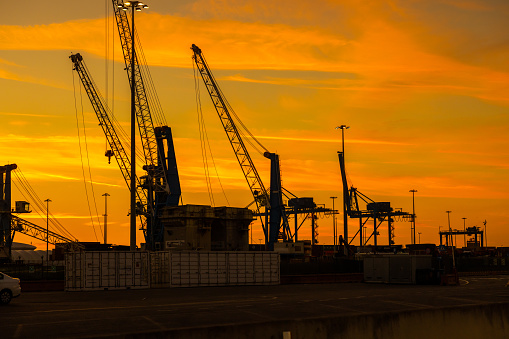Tall cranes and cargo containers by light poles at commercial dock in front of orange sky during sunset
