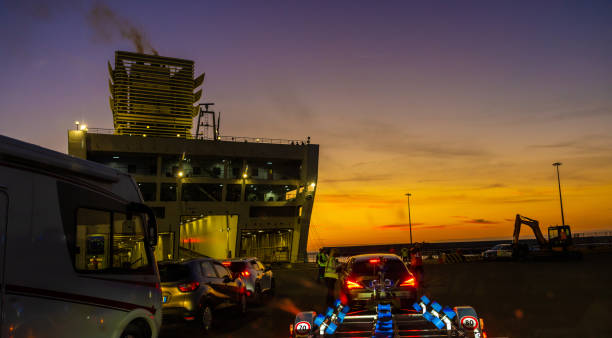 Cars and trucks in row moving towards illuminated ship at harbor with orange sunset sky in background
