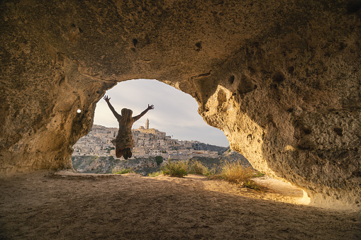 Rear view of carefree woman jumping with arms raised in cave on sunny day during vacation
