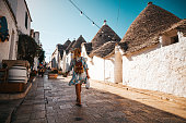 Rear view of female tourist with backpack walking in alley amidst trulli houses in a row against clear sky at Alberobello