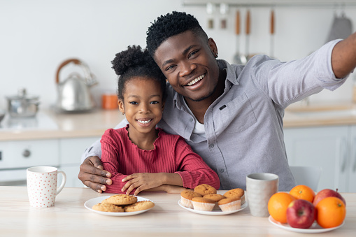 Happy black family single father and little daughter taking selfie while having breakfast together at home, sitting at kitchen table, embracing and smiling at camera, copy space