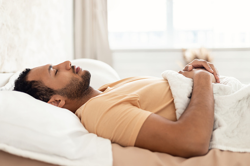 Arabic Young Man Sleeping Lying On Back Holding Hands On Stomach Resting In Bed In Modern Bedroom Indoor. Guy Napping At Home In The Morning. Comfort And Recreation Concept