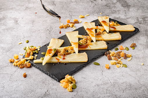 From above of appetizing sliced cheese served on black board with nuts and dried fruits placed on gray tabletop and sprinkled with honey from spoon