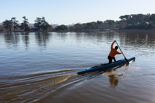 Anonymous tourist standing on kayak and rowing in calm rippling lake against trees in countryside