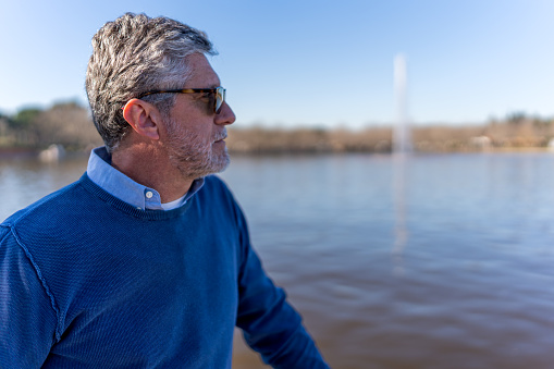 Thoughtful elderly male in sunglasses and casual clothes standing on shore of lake while admiring picturesque scenery on sunny day
