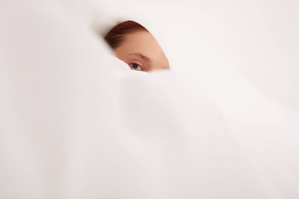 Young woman with only one eye visible through a hole in white fabric.