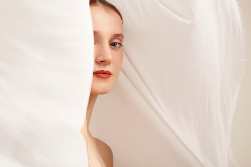 Portrait of young woman emotionally looking at camera through flowing white fabric.