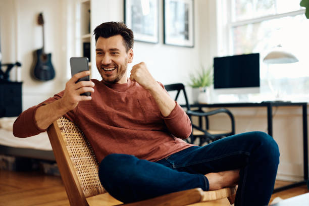 Cheerful man celebrating good news while using mobile phone at home. Happy man receiving good news while reading text message on cell phone at home. sports betting stock pictures, royalty-free photos & images