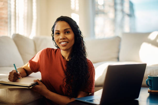 istock Happy black woman using laptop and writing notes while studying at home and looking at camera. 1466797094