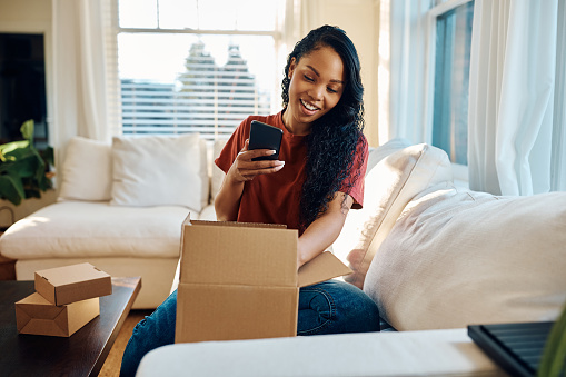 Happy African American woman using mobile phone while unpacking delivered cardboard box at home.