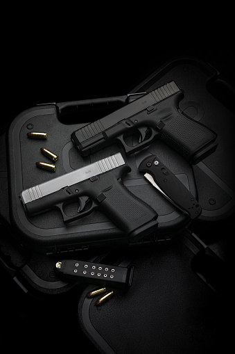 Two pistols, 9mm cartridges and a penknife lie on a plastic gun case. Self-defense and survival kit. Compact edged weapons and firearms. Dark background.