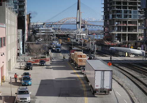 New Westminster, Canada - February 14, 2023: Pedestrians, workers and drivers share the busy level crossing on Front Street and Begbie Street. Background shows the cable-stayed TransLink SkyBridge and the orange Pattullo Bridge over the Fraser River. Winter morning in Metro Vancouver.