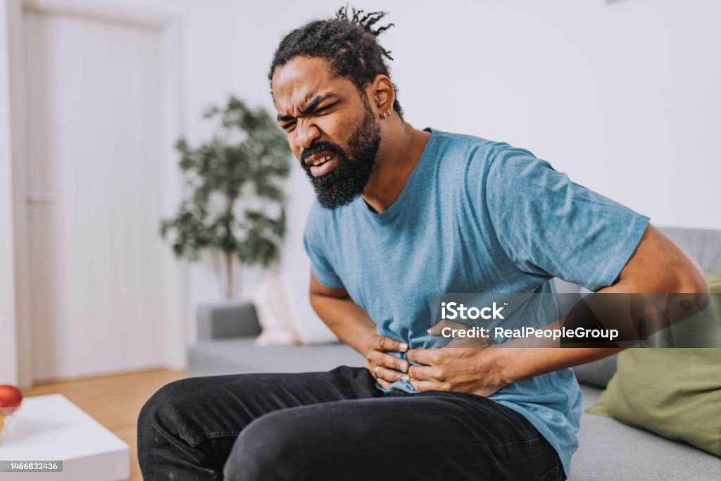 Man with a stomachache A mid-adult African-American man is sitting in his living room and holding his painful stomach. Irritable Bowel Syndrome Stock Photo