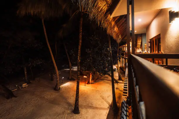 Top view in a low key and long exposure from the balcony of a straw-roofed hotel to the entrance of the property with white sand, tall palm trees, and trees enriching the landscape on a summer night