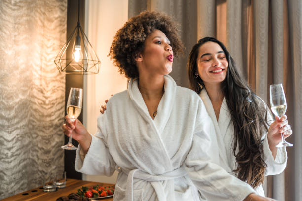 Two Cheerful Diverse Friends In Bathrobes Drinking Wine And Eating Cupcake stock photo