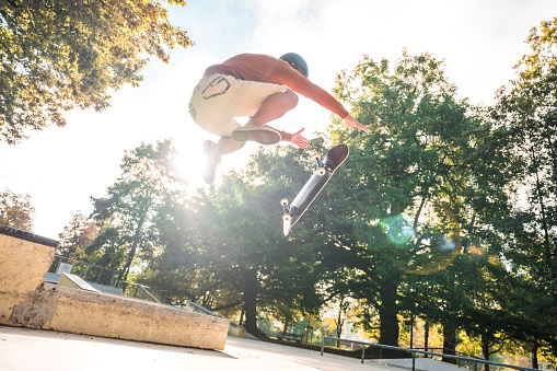 Male skateboarder performing a high jump while wearing protective helmet. Full length shot, sunbeam coming from behind him. Trees in the background.