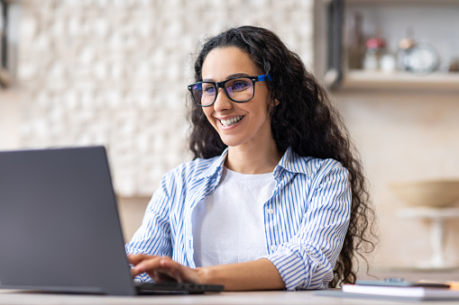 Positive latin female freelancer in eyeglasses using laptop at home, sitting at table in kitchen, typing on keyboard and smiling. Remote job concept