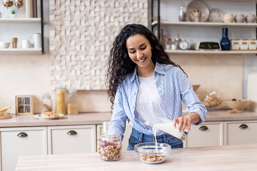 Breakfast concept. Happy latin woman pouring fresh milk from glass into bowl with cereals, having delicious meal in kitchen interior, copy space