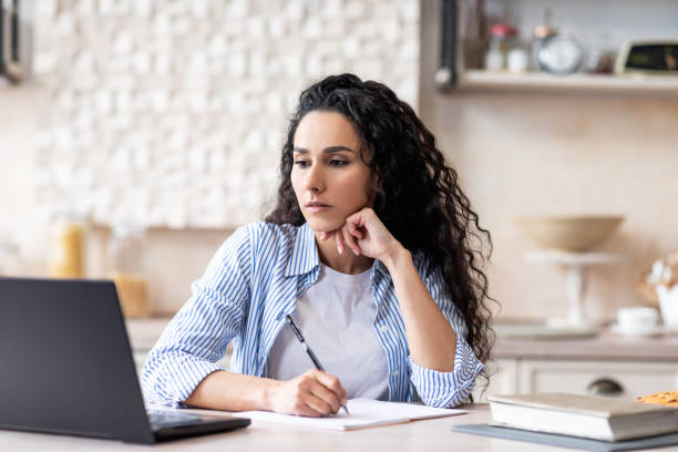 istock Latin lady studying remotely from home, looking at laptop and making notes, sitting at table in kitchen interior 1466839554