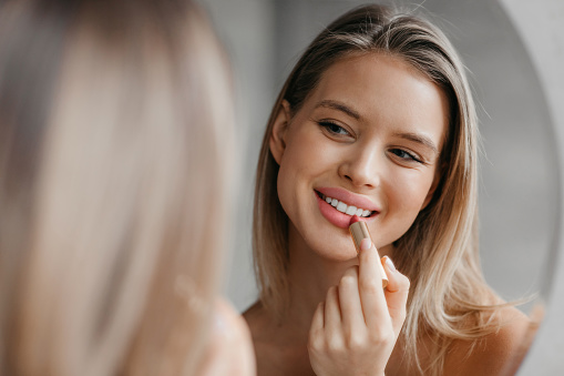 Routine procedures and decorative cosmetics for homemade visage. Happy young woman applying lipstick in bathroom