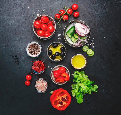 Vegetables, olives, oil and ingredients for cooking greek salad with feta cheese, cherry tomato, paprika, cucumber and red onion, healthy mediterranean food, low calories eating. Black stone background, top view