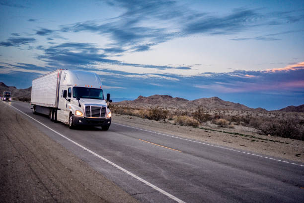 White Semi-Truck Traveling Down a Two-Lane Highway at Dusk Under a Beautiful Cloudscape in a Hilly Desert Area Southwest United States With Headlights On stock photo