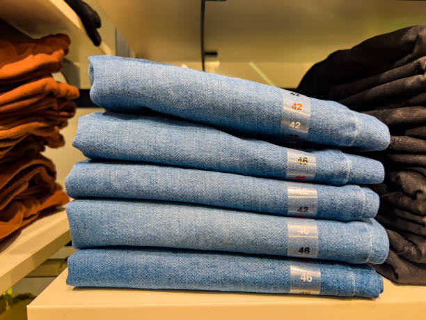 Row of blue Jeans in fashion industry. Tidy stack of folded trousers in a retail store envionment with various size number labels. Casual business cloth. Stone washed, sandpbaster or weathered blue cotton textile. Blue manufactured object backgrond, copy space.