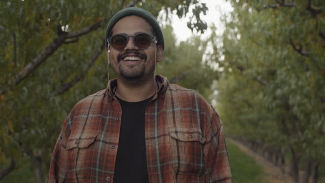 Hand Held Video Portrait Shot of An Hispanic Man In His Thirties Wearing Sun Glasses And A Beard Wearing A Beanie Hat And Red And Orange Flannel In A Peach Orchard With Tons Of Leaves Looks Straight At Camera And Then Gives The Hang Loose Hand Signal