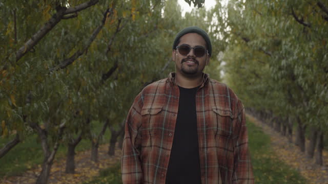 Hand Held Video Portrait Shot of An Hispanic Man In His Thirties Wearing Sun Glasses And A Beard Wearing A Beanie Hat And Red And Orange Flannel In A Peach Orchard With Tons Of Leaves Looks Straight At Camera And Then Gives The Peace Sign Hand Signal