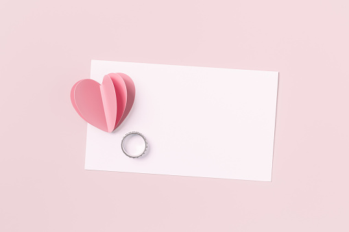 Pink paper heart and empty white paper card and wedding ring on pink colored background. Minimal style love note, pastel color, mock up valentine card, marriage proposal. Romantic concept, top view