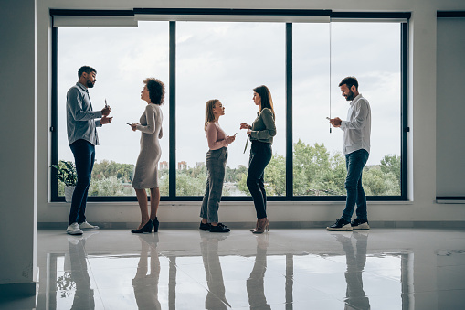 Shot of a business persons standing side by side against the window in modern office. Shot of a five confident young businesspeople standing next to each other and sharing business ideas. Multi-ethnic group of business persons in business meeting or business seminar. Their reflection is seen in the floor.