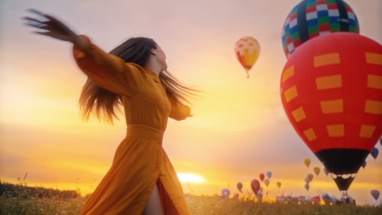 Carefree young woman spinning in sunrise field with hot air balloons