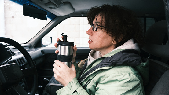 Mature woman in the car is drinking from travel mug
