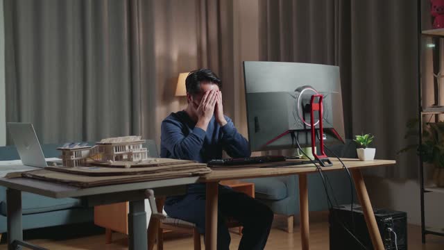 Asian Male Engineer With The House Model Having A Headache While Working On A Desktop At Home